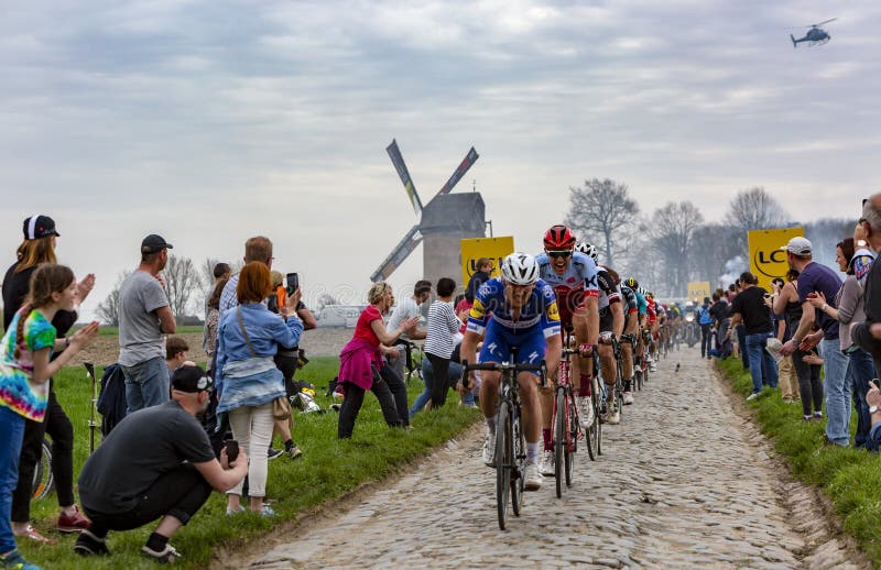 Templeuve, France - April 08, 2018: The peloton riding on the cobblestone road in Templeuve in front of the traditional Vertain Windmill during Paris-Roubaix 2018. Templeuve, France - April 08, 2018: The peloton riding on the cobblestone road in Templeuve in front of the traditional Vertain Windmill during Paris-Roubaix 2018