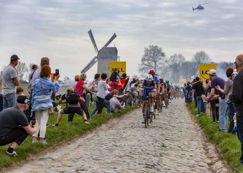 Templeuve, France - April 08, 2018: The peloton riding on the cobblestone road in Templeuve in front of the traditional Vertain Windmill during Paris-Roubaix 2018. Templeuve, France - April 08, 2018: The peloton riding on the cobblestone road in Templeuve in front of the traditional Vertain Windmill during Paris-Roubaix 2018