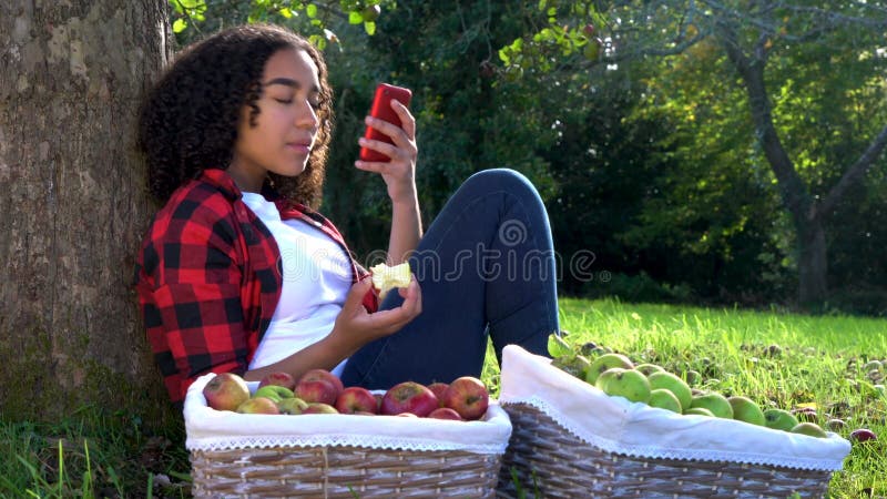 Penchement se reposant de jeune femme d'adolescente contre un arbre dans un verger mangeant une pomme utilisant un téléphone port clips vidéos