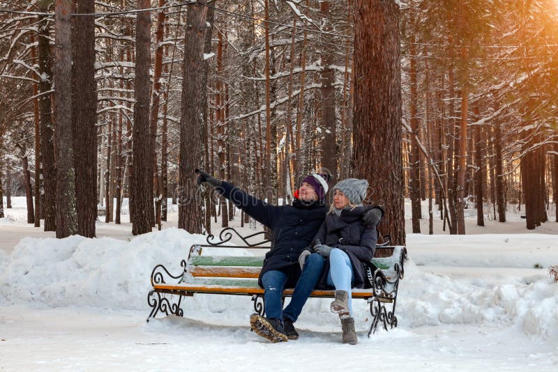 Pendant l'après-midi d'hiver, un couple affectueux dans des vestes et les chapeaux se reposent sur un banc dans les bois dans la photos libres de droits Pendant l'après-midi d'hiver, un couple affectueux dans des vestes et les chapeaux se reposent sur un banc dans les bois dans la photos libres de droits