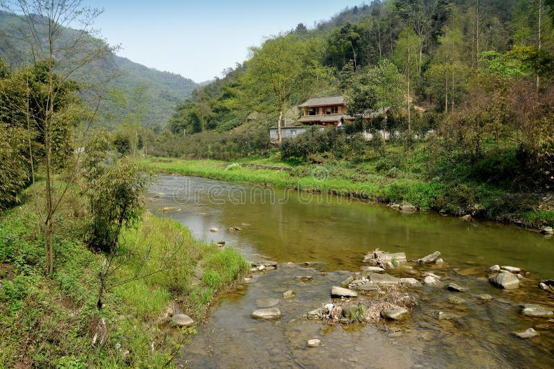 An old wooden Sichuan style farmhouse nestles along the shoreline of a clear-water mountain river coursing through a lush valley and hillsides covered with bamboo and various deciduous trees in Pengzhou, China. An old wooden Sichuan style farmhouse nestles along the shoreline of a clear-water mountain river coursing through a lush valley and hillsides covered with bamboo and various deciduous trees in Pengzhou, China.