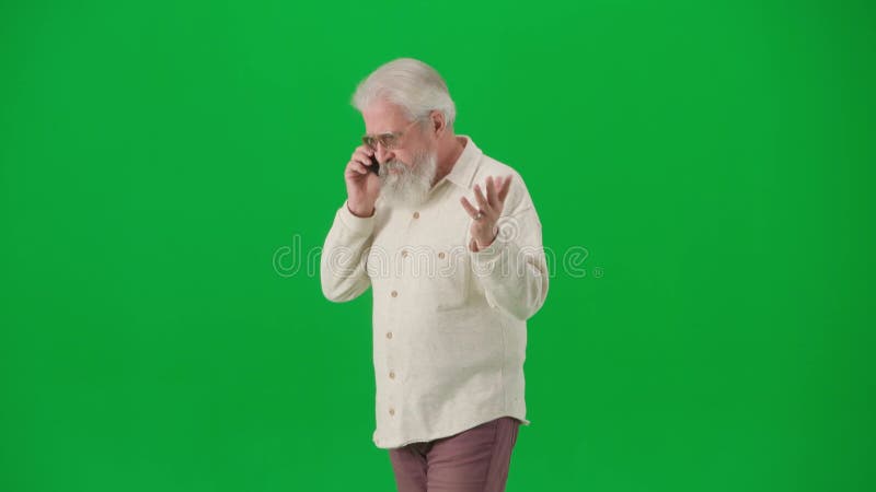 Portrait of aged bearded man on chroma key green screen. Senior man walking and talks on smartphone. Half turn..