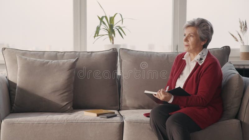Pensionerad kvinna som sitter på soffan in i vardagsrum med det stora fönstret och skriver dagboken