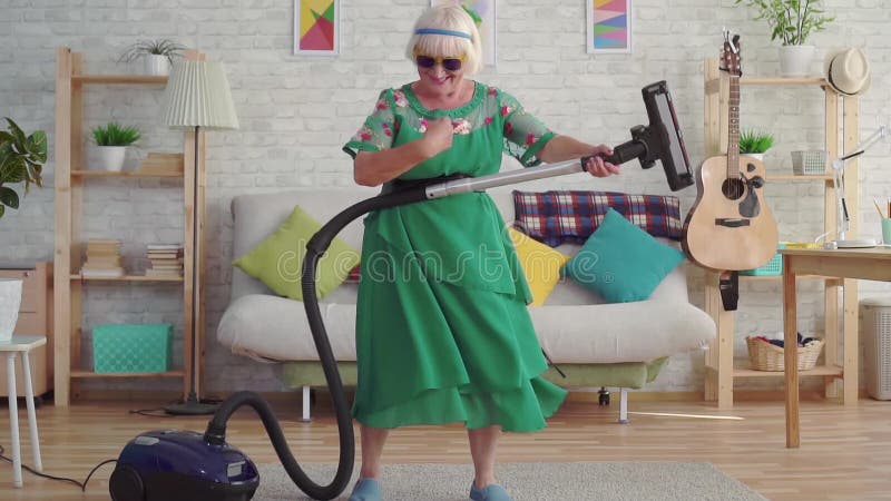 Pensionista feliz alegre de la mujer mayor con el pelo gris en los vidrios que juegan un aspirador como una guitarra MES lento almacen de metraje de vídeo
