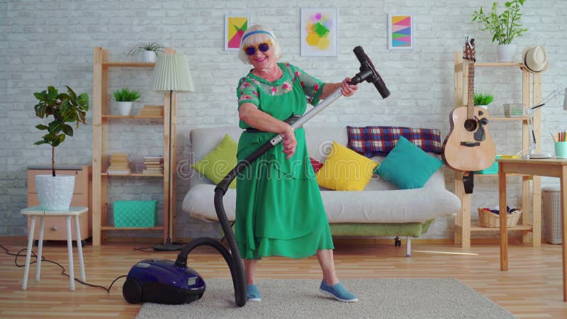 Pensionista feliz alegre de la mujer mayor con el pelo gris en los vidrios que juegan un aspirador como una guitarra almacen de metraje de vídeo