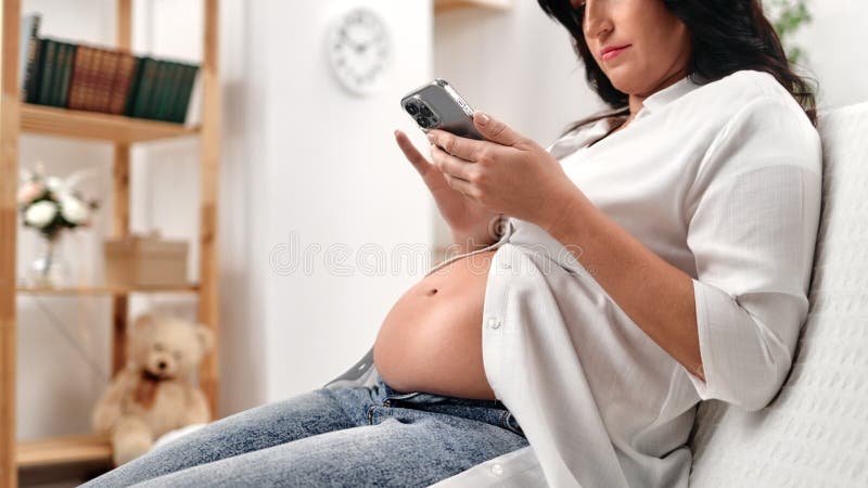 Pensische brunette zwangere vrouw met buik gebruikt smartphone die ontspannen is op bank bij thuissluiting