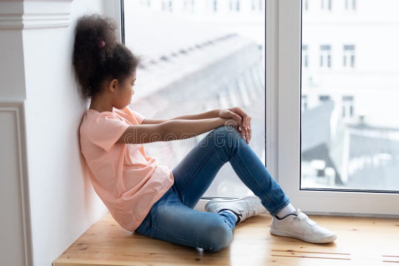 Pensive african american young girl sitting alone on windowsill.