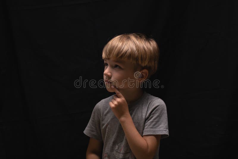 Pensive beau petit garçon blonde avec doigt près de la bouche sur fond noir.