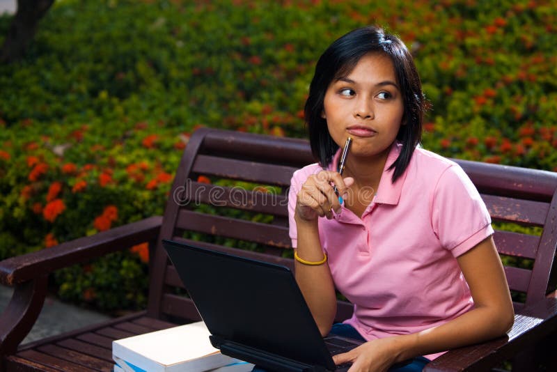 A cute pensive Asian college student thinks while using her laptop on a university campus bench. 20s female Asian Thai model of Chinese descent. A cute pensive Asian college student thinks while using her laptop on a university campus bench. 20s female Asian Thai model of Chinese descent.