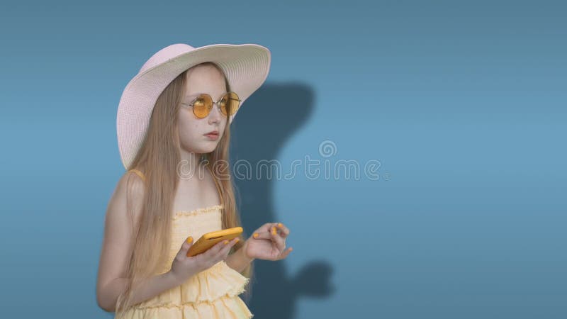 Pensive teenager girl looking up and typing text on smartphone on blue background. Thinking young girl using mobile and stock video