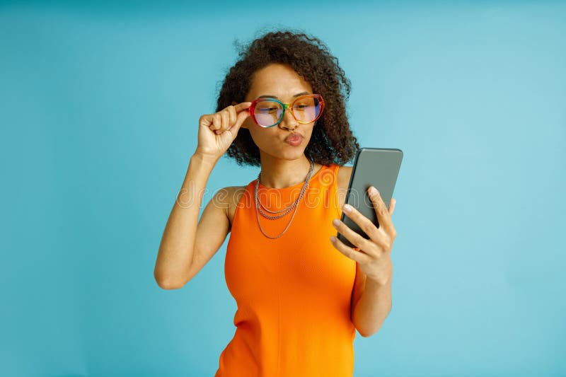 Pensive woman in eyeglasses looking on mobile phone while standing on blue studio background