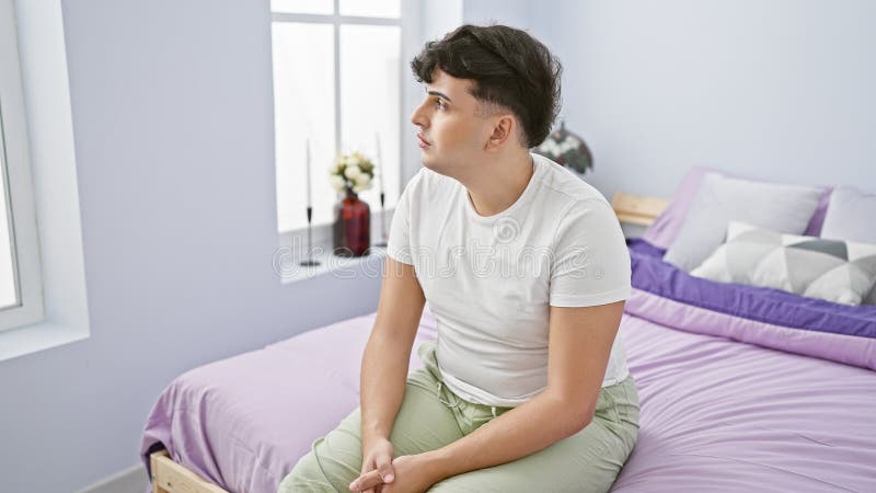 Pensive young man sitting on a bed in a bright, modern bedroom, looking away thoughtfully royalty free stock photos