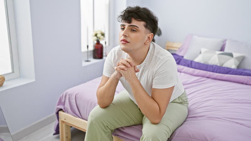 Pensive young man sitting on bed in a modern bedroom, with a contemplative expression and casual clothing royalty free stock images