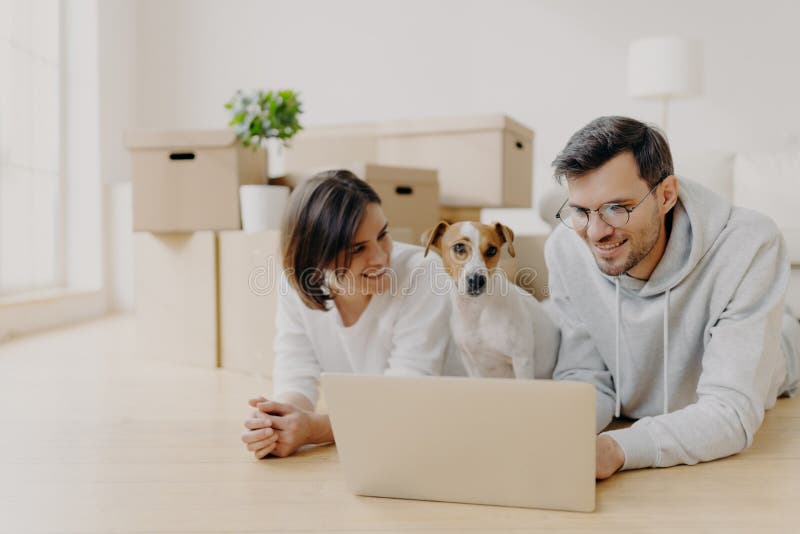 People and new apartment concept. Happy young husband and wife relax on floor, use modern laptop computer for surfing net, pose in