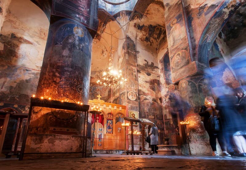 People praying with candles in dark interior of Church of the Archangels with frescoes