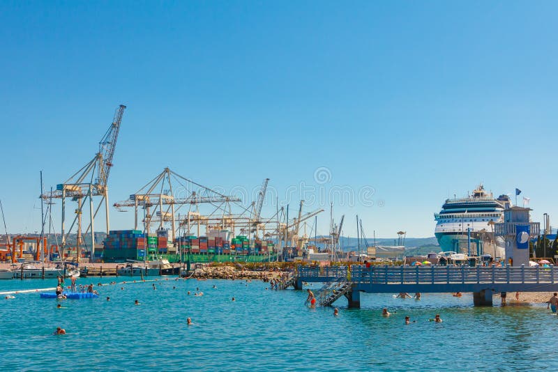 People swimming on the beach in front of harbor cranes and luxury cruise liner. Koper, Slovenia