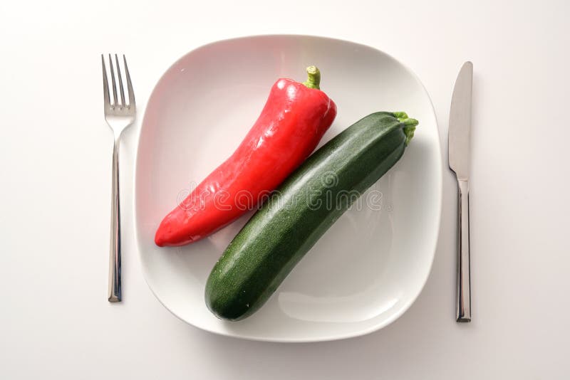 Raw red pointed pepper and zucchini on a white plate and cutlery on a bright background, healthy diet with mediterranean vegetables to lose weight in the new year, copy space, high angle view from above. Raw red pointed pepper and zucchini on a white plate and cutlery on a bright background, healthy diet with mediterranean vegetables to lose weight in the new year, copy space, high angle view from above