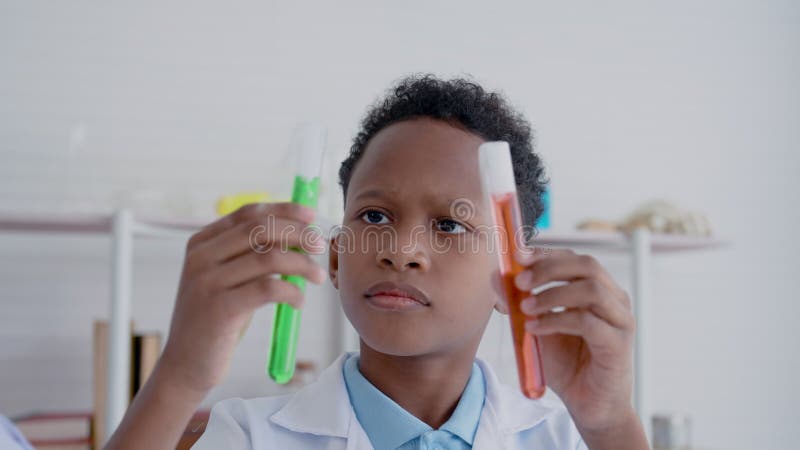 Pequeno garoto asiático de 4k especula e se pergunta líquido colorido em um tubo de ensaio em ambas as mãos em uma escola primária