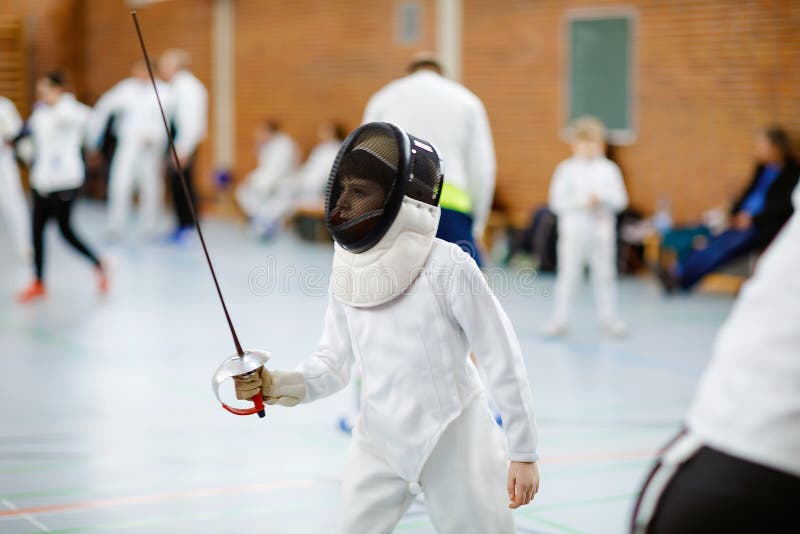 Pequeno garoto esgrimindo na cerca Criança com uniforme de encanador branco com máscara e sabre Formação de crianças ativas fotografia de stock royalty free Pequeno garoto esgrimindo na cerca Criança com uniforme de encanador branco com máscara e sabre Formação de crianças ativas fotografia de stock royalty free