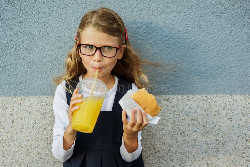 Pequeña colegiala sonriente linda que sostiene una hamburguesa y una naranja imagenes de archivo Pequeña colegiala sonriente linda que sostiene una hamburguesa y una naranja imagenes de archivo