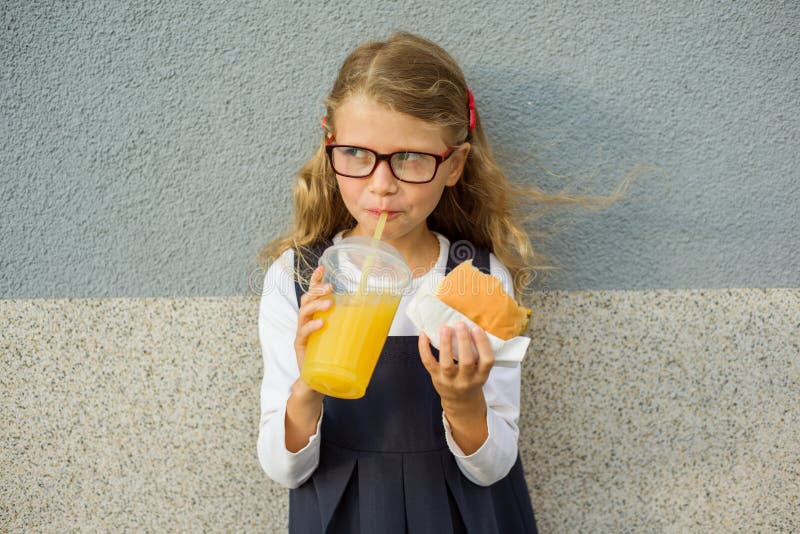 Pequeña colegiala sonriente linda que sostiene una hamburguesa y una naranja foto de archivo Pequeña colegiala sonriente linda que sostiene una hamburguesa y una naranja foto de archivo