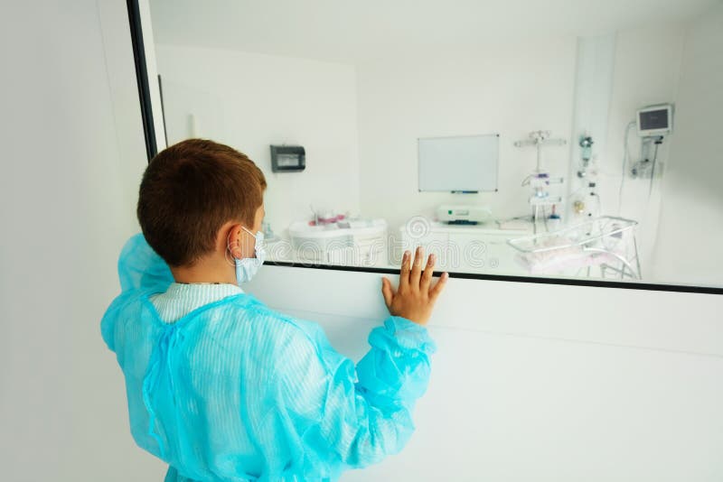 Little boy near the window of ICU room in hospital wearing medical mask and robe. Little boy near the window of ICU room in hospital wearing medical mask and robe