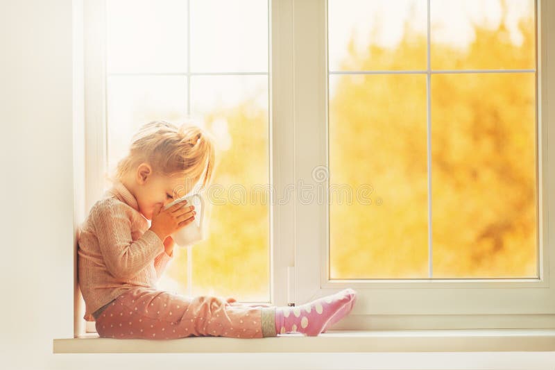 Pequeña muchacha linda del niño que se sienta por la taza que se sostiene interior de la ventana de cacao caliente de la bebida q fotos de archivo libres de regalías Pequeña muchacha linda del niño que se sienta por la taza que se sostiene interior de la ventana de cacao caliente de la bebida q fotos de archivo libres de regalías