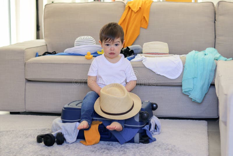 Pequeño bebé lindo con el sombrero divertido de la tenencia de la expresión de la cara en las manos que se sientan en la maleta l imagen de archivo libre de regalías Pequeño bebé lindo con el sombrero divertido de la tenencia de la expresión de la cara en las manos que se sientan en la maleta l imagen de archivo libre de regalías