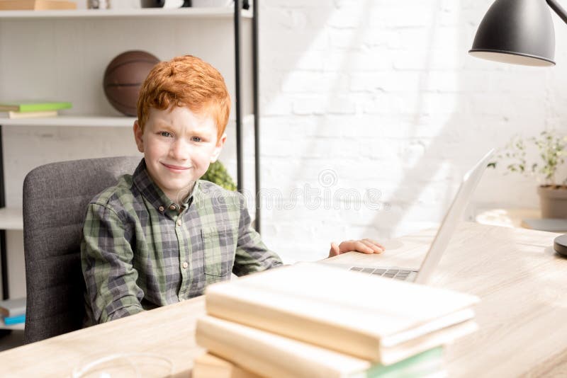 Pequeño muchacho lindo del pelirrojo que sonríe en la cámara mientras que se sienta en la tabla con los libros foto de archivo libre de regalías
