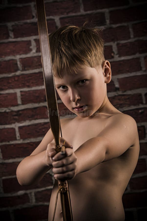 Blond hair boy drawing a bow and arrow. Looking straight to the camera. Red brick wall background. Blond hair boy drawing a bow and arrow. Looking straight to the camera. Red brick wall background.