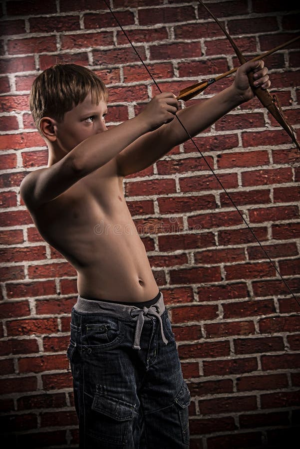 Blond hair boy drawing a bow and arrow. Looking up. Red brick wall background. Blond hair boy drawing a bow and arrow. Looking up. Red brick wall background.