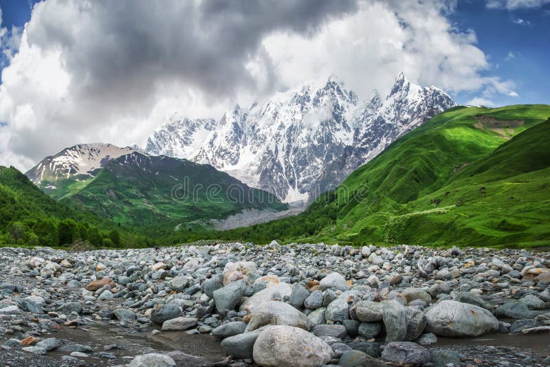 Perfect mountain landscape in Georgia, Svaneti. Green hills, snowy rocks mounts and stones on river bank against sky with clouds