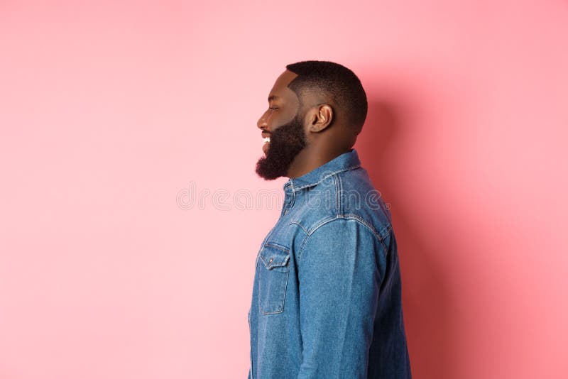 Perfil de un guapo negro barbudo parado sobre fondo rosado sonriendo y mirando a la izquierda en el espacio de copia imagenes de archivo