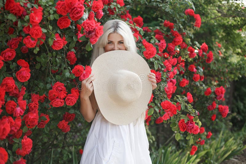Young beautiful woman in a hat, near a large bush of red roses in the spring garden outdoors