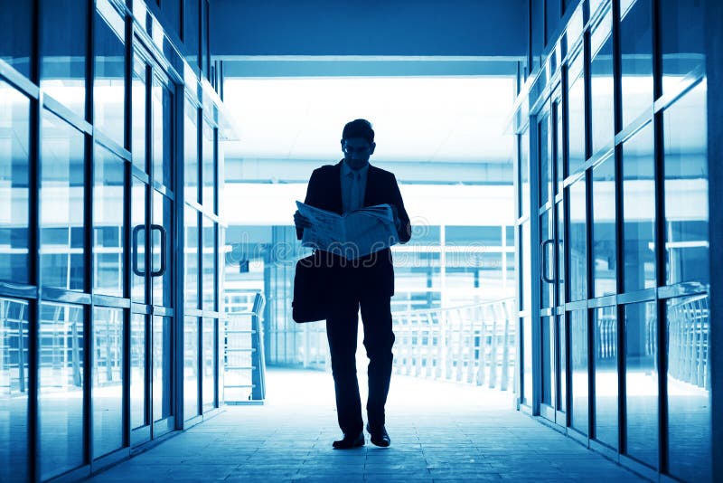 Silhouette of business man residing newspaper and walking thru modern office building, in blue tone. Silhouette of business man residing newspaper and walking thru modern office building, in blue tone.