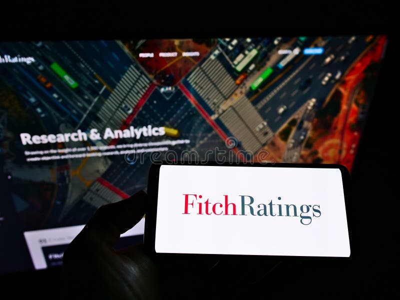 Person holding mobile phone with logo of American credit rating agency Fitch Ratings Inc. on screen in front of web page.