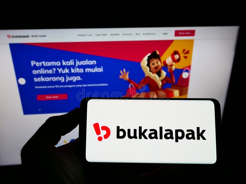 Person holding mobile phone with logo of Indonesian e-commerce company Bukalapak on screen in front of business web page.
