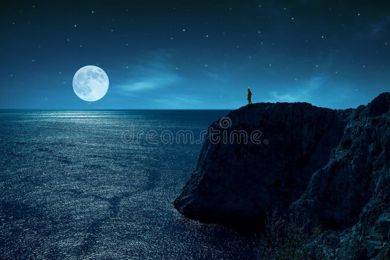 Person is standing in the edge of a cliff against the sea and full moon, under stars and moon light. stock photo Person is standing in the edge of a cliff against the sea and full moon, under stars and moon light. stock photo