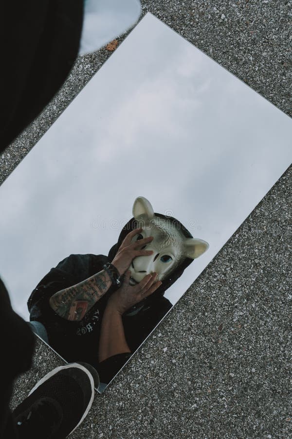 Person with tattoos wearing a sheep mask and black hoodie looking through a mirror on the ground stock photo Person with tattoos wearing a sheep mask and black hoodie looking through a mirror on the ground stock photo
