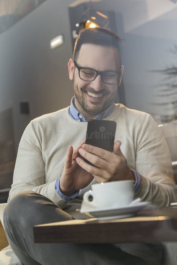 Portrait of a handsome young man sitting at a restaurant table, typing a text message on a smart phone and drinking coffee. Portrait of a handsome young man sitting at a restaurant table, typing a text message on a smart phone and drinking coffee