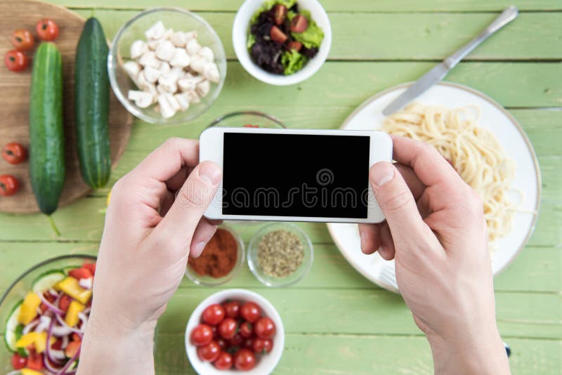 Persona que sostiene smartphone con la pantalla en blanco y que fotografía los espaguetis y las verduras frescas en la tabla de m imágenes de archivo libres de regalías Persona que sostiene smartphone con la pantalla en blanco y que fotografía los espaguetis y las verduras frescas en la tabla de m imágenes de archivo libres de regalías