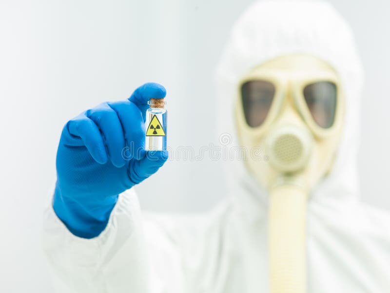 Person in bio hazard suit and gas mask holding a radioactive isotope sample in a transparent colorless vial. Person in bio hazard suit and gas mask holding a radioactive isotope sample in a transparent colorless vial