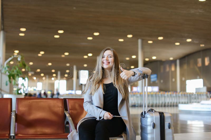 Personne féminine heureuse s'asseyant dans le hall d'aéroport avec le comprimé moderne près de la mallette et montrant des pouces photos stock Personne féminine heureuse s'asseyant dans le hall d'aéroport avec le comprimé moderne près de la mallette et montrant des pouces photos stock
