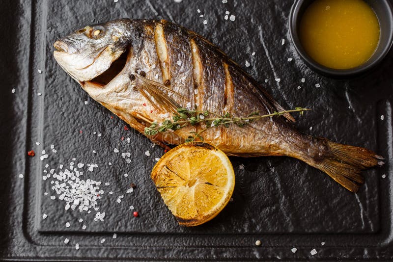 Pescados asados a la parrilla deliciosos de la brema del dorado o de mar con las rebanadas del limón, especias, romero en piedra imagen de archivo Pescados asados a la parrilla deliciosos de la brema del dorado o de mar con las rebanadas del limón, especias, romero en piedra imagen de archivo