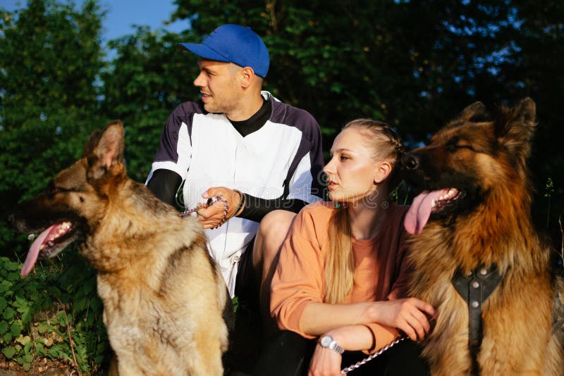 Pet Love, Dog Training, best dog breeds for family. Young sports couples walking with two German Shepherd dogs outdoors in summer park. Pet Love, Dog Training, best dog breeds for family. Young sports couples walking with two German Shepherd dogs outdoors in summer park.