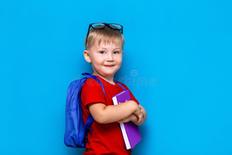 Petit garçon de sourire heureux avec les verres sur sa tête, livre dans des mains, cartable sur ses épaules De nouveau ? l'?cole photographie stock libre de droits Petit garçon de sourire heureux avec les verres sur sa tête, livre dans des mains, cartable sur ses épaules De nouveau ? l'?cole photographie stock libre de droits