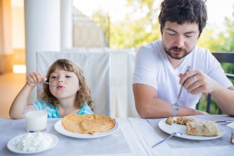 Petite fille adorable prenant le petit déjeuner avec son père, mangeant des crêpes et du lait boisson image stock Petite fille adorable prenant le petit déjeuner avec son père, mangeant des crêpes et du lait boisson image stock