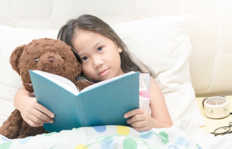 Petite fille asiatique mignonne dans la chemise de nuit lisant un livre avec le nounours b photographie stock Petite fille asiatique mignonne dans la chemise de nuit lisant un livre avec le nounours b photographie stock
