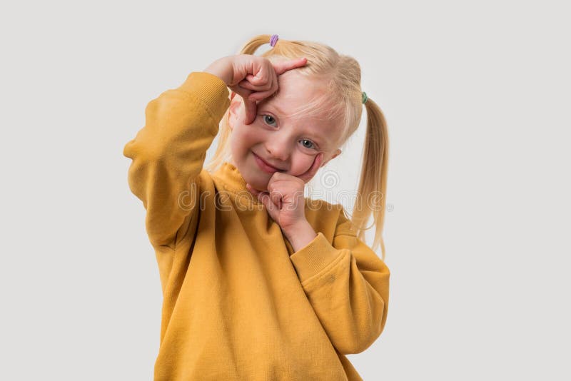 Petite fille aux cheveux blonds dans un pull jaune tient sa tête avec ses mains et regarde la caméra images libres de droits