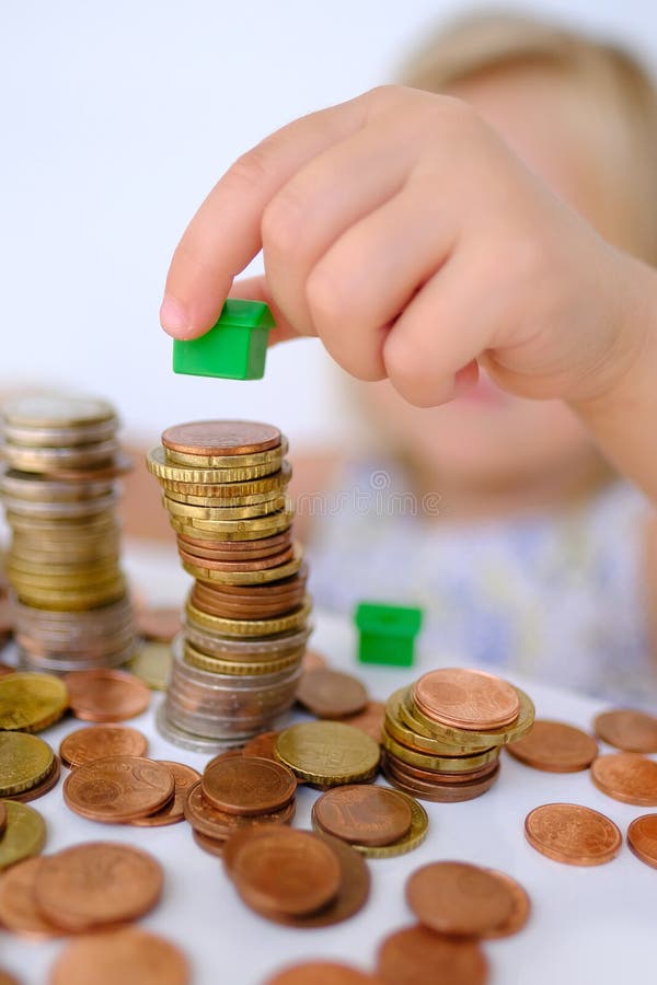 Small child, blonde girl 3 years old playing with cash, house model on stacks euro currency coins, pocket money, concept home mortgage, home insurance, financial literacy of children, personal savings. Small child, blonde girl 3 years old playing with cash, house model on stacks euro currency coins, pocket money, concept home mortgage, home insurance, financial literacy of children, personal savings